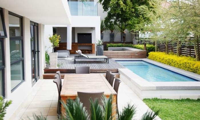 Área externa residencial com mesas, cadeiras, palmeiras, vasos de plantas e arbustos e árvores ao redor de uma piscina pequena.